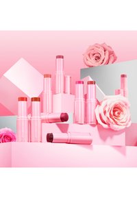 Eight pink lipstick sticks with a glossy finish, displayed on pink geometric blocks, accompanied by light pink roses.