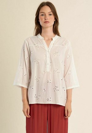 Giovane donna che indossa una camicetta bianca in sangallo con maniche a 3/4 e pantaloni rossi a gamba larga, in piedi davanti a uno sfondo beige uniforme.