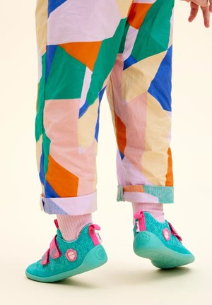 Scarpe per bambini color turchese con cinturini in velcro rosa e una superficie testurizzata, abbinate a pantaloni con un motivo geometrico multicolore.