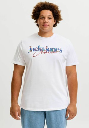 Młody mężczyzna z kręconymi włosami, ubrany w białą koszulkę Jack & Jones Denim i jasnoniebieskie jeansy na jednolitym, jasnym tle.