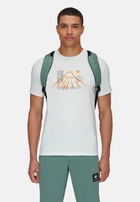 Vit t-shirt med grafik av ett berg och text. Kortsleeves, rund hals, parat med tealbyxor och en grön ryggsäck.