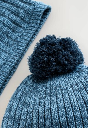 Strickmütze in verschiedenen Blautönen, mit einem flauschigen dunkelblauen Pompon oben drauf. Ein Schal aus ähnlichem Material und in ähnlicher Farbe ist teilweise sichtbar.