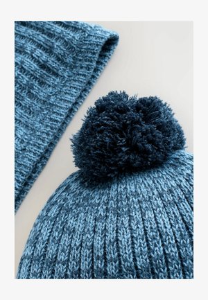 Adi cepure dažādās zilās nokrāsās ar pūkainu tumši zilu pom-pomu augšpusē. Šalle, kas izgatavota no līdzīgas tekstūras materiāla un krāsas, ir daļēji redzama.