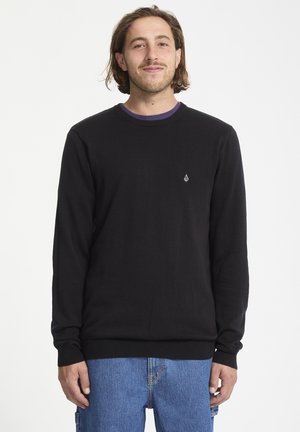 Zwarte gebreide trui met een ronde hals en lange mouwen, met een klein logo op de linkerborst. Gecombineerd met blauwe denim jeans.