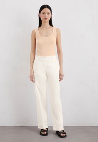 Breskva majica bez rukava sa širokim izrezom, uparena sa bijelim hlačama ravnog kroja. Outfit je upotpunjen crnim sandalama i jednostavnom, glatkom teksturom.