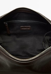 De binnenkant van een leren bruine tas met een zwarte stoffen voering, een ritsvak en een label. De rits is zilverkleurig met een gladde afwerking.