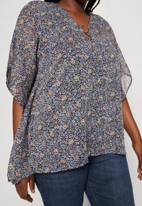 Blusa floreale blu navy con maniche corte a farfalla, dotata di scollatura a V e dettaglio a cerchio decorativo sul colletto. Tessuto leggero e trasparente.