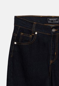 Jeans de mezclilla azul oscuro con costuras amarillas, detalle de bolsillo delantero y cierres de botón plateado. La etiqueta dice "Blue Effect, talla 11-12 años."