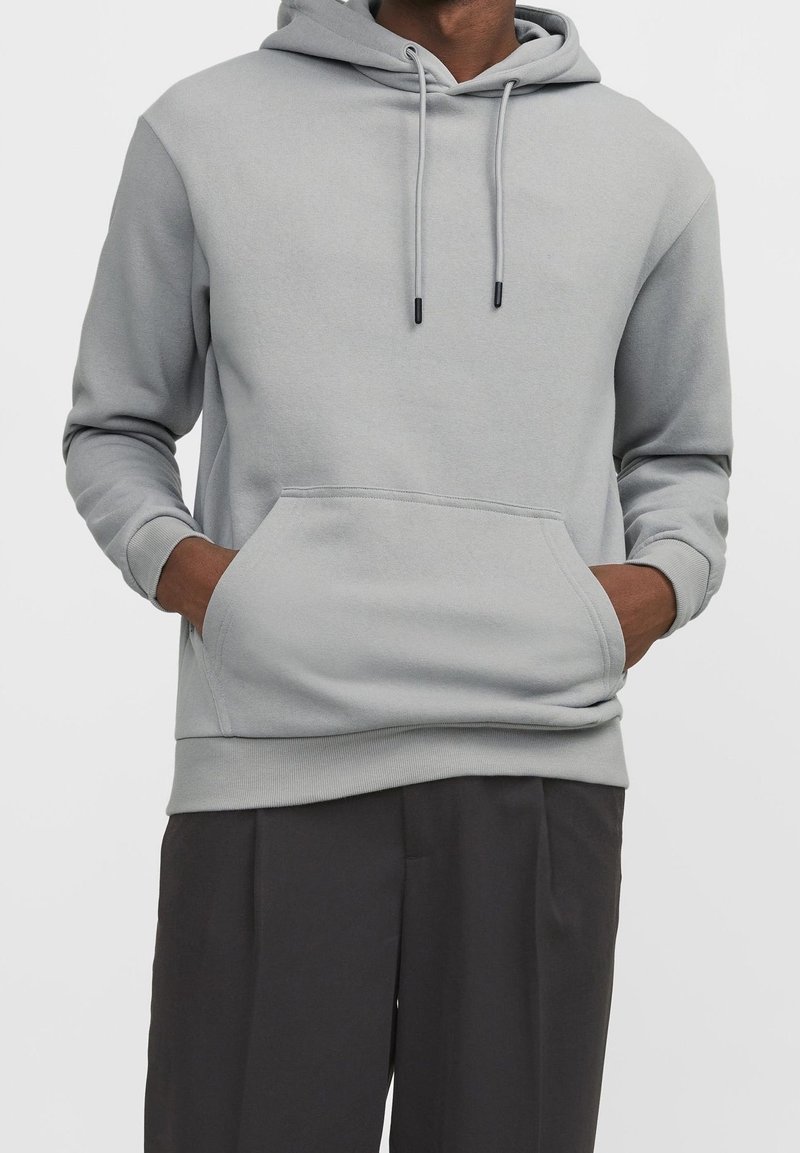 Homme portant un sweat à capuche gris clair avec poche avant et cordons, associé à un pantalon sombre et ample, mains dans les poches.