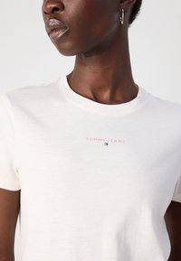 Balta medvilninė marškinė su trumpomis rankovėmis, viduryje esantis rožinis „TOMMY JEANS“ logotipas ir po juo mažas spalvotas emblema.