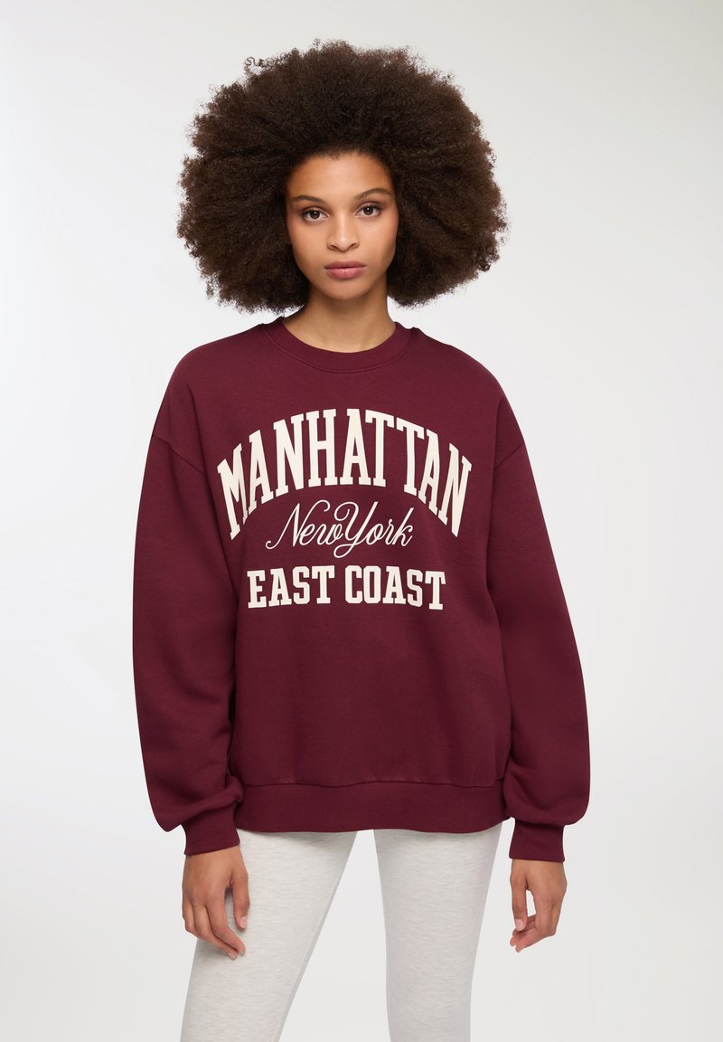 Felpa bordeaux con la scritta "MANHATTAN New York EAST COAST" stampata in crema. Maniche lunghe, polsini a costine e texture morbida.
