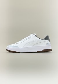 Λευκό sneaker Puma χαμηλού τύπου με γκρι ταμπέλα στη φτέρνα, λευκά κορδόνια, διάτρητες πλευρές και καφέ λαστιχένια σόλα σε ουδέτερο φόντο.