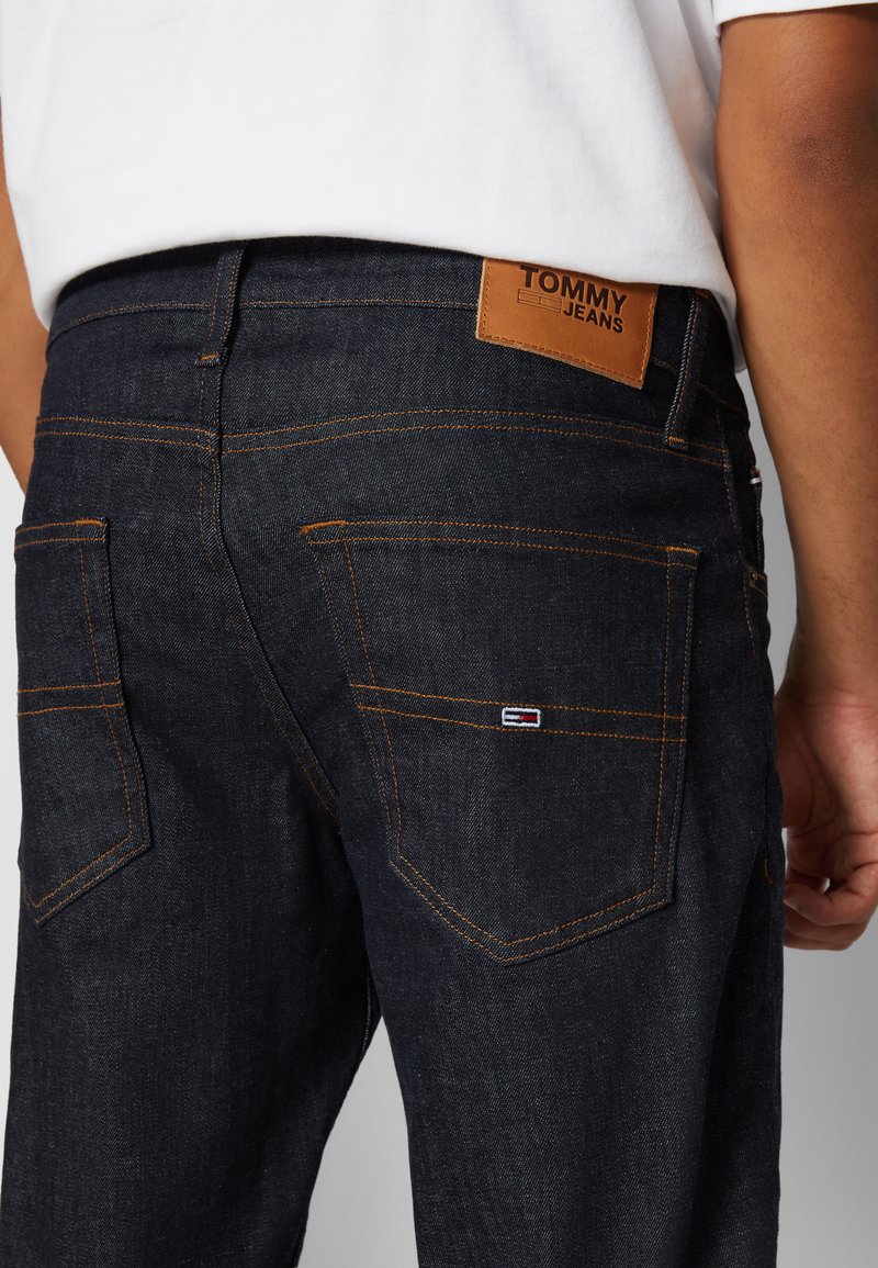 Donkerblauwe denim jeans met oranje stiksels, met een leren label "TOMMY JEANS" op de achterkant van de tailleband. Twee achterkantzakken zichtbaar.