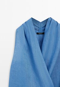Blå linnelångärmad omlottopp med djup V-ringning och vikta detaljer runt kragen mot vit bakgrund.