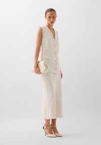 Gilet senza maniche di colore crema intrecciato con bottoni frontali, abbinato a una gonna a fantasia coordinata e scarpe con tacco crema. Tiene una borsa a tracolla leggera.