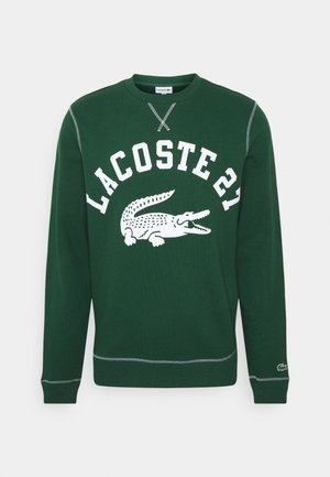 Ciemnozielona bluza crewneck z białym napisem "LACOSTE 21" i logo krokodyla na środku, ściągaczami przy rękawach i u dołu z kontrastowym obszyciem.