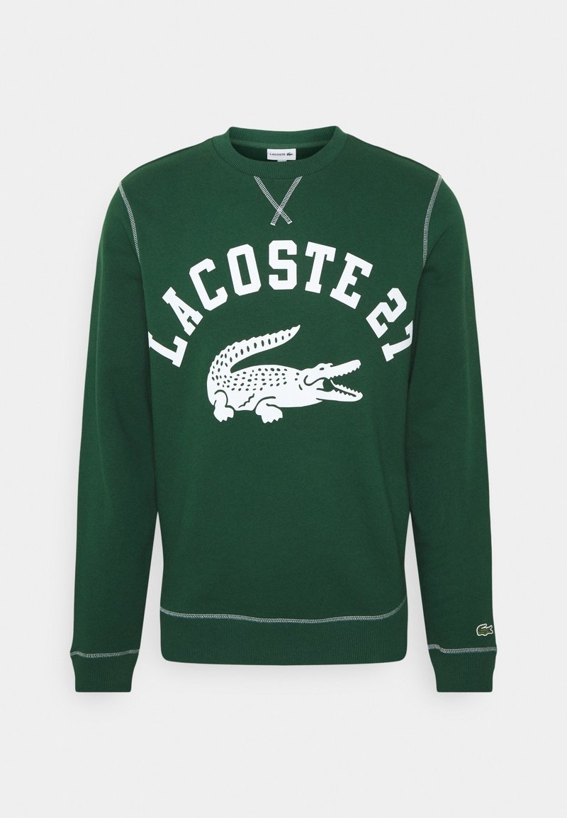 Sweat-shirt vert foncé à col rond avec le texte blanc "LACOSTE 21" et le logo crocodile centrés, poignets et ourlet côtelés avec surpiqûres contrastées.