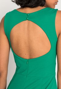 Vestido verde com um decote alto, recorte nas costas e dois botões decorativos. O tecido parece suave, com um ligeiro brilho.