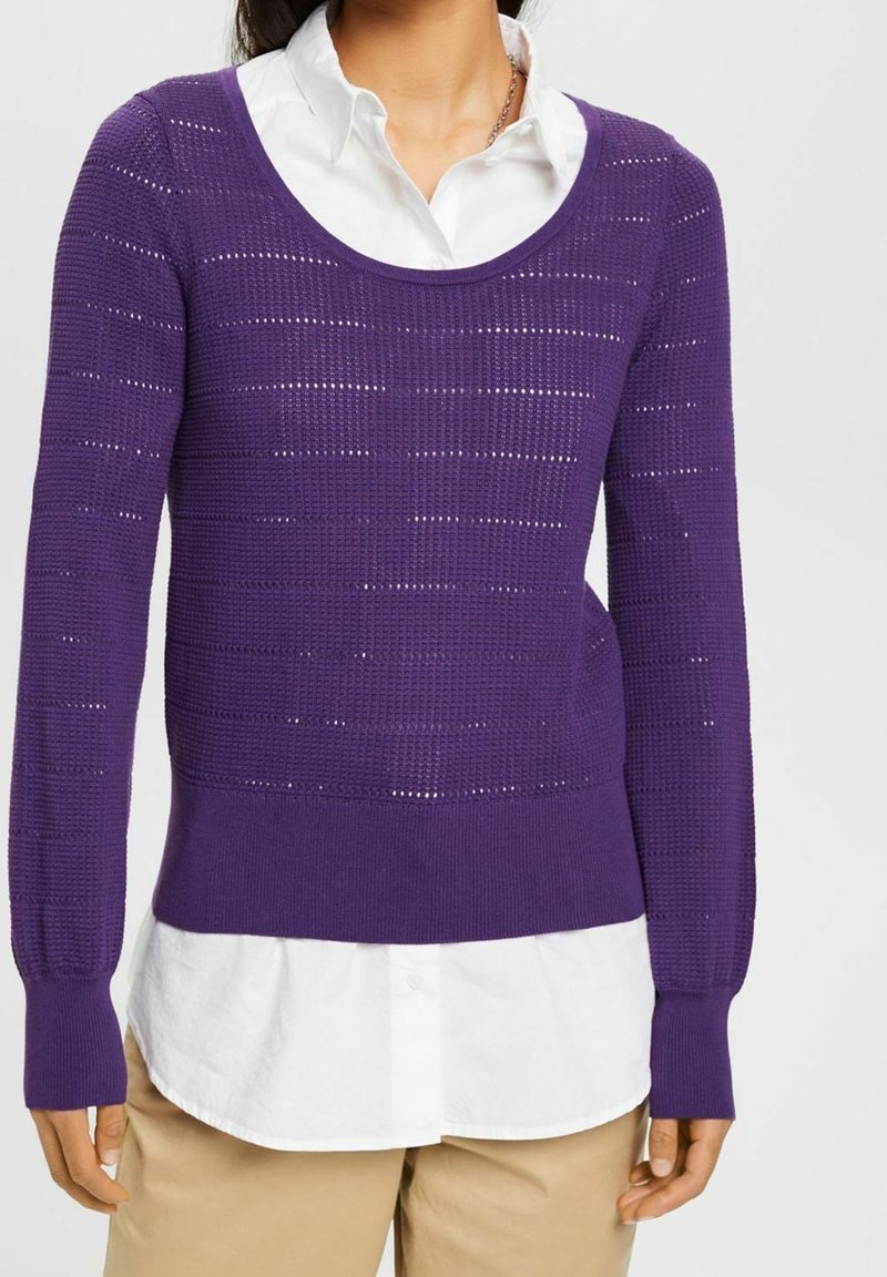 Femme portant un pull en maille violet par-dessus une chemise blanche à col, et un pantalon beige, debout devant un fond clair uni.