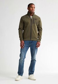 Olivově zelená zipová bunda s vysokým límcem, strukturovanou tkaninou a kapsou na prsou. Nositelná s modrými džíny a bílými teniskami.