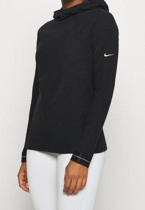 Frau trägt ein strukturiertes schwarzes Langarm-Hoodie mit weißem Nike-Logo auf dem linken Ärmel und weiße Hose.