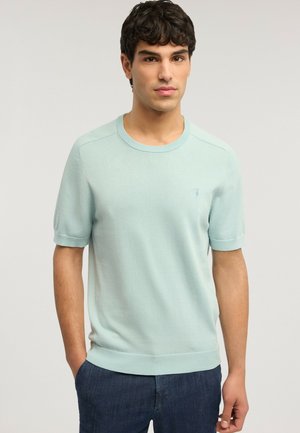 Ung mand med mørkt hår iført en lyseblå kortærmet striktrøje og mørkeblå bukser, stående med hænder delvist i lommerne.