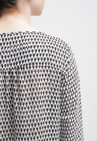 Svart och vit mönstrad blus med en scallop-design, genomskinligt tyg och rund halsringning med detaljer på den samlade baksidan.
