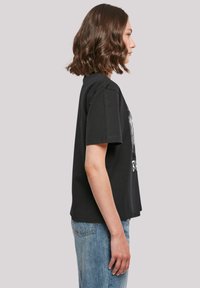 Black cotton t-shirt with short sleeves, loose fit, featuring a graphic print on the front. Paired with light blue denim jeans.