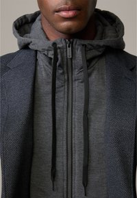 Szary rozpinany sweatshirt z teksturowanym wzorem, z czarnymi sznurkami i ciepłym kapturem, w połączeniu z ciemnym blazerm we wzory.