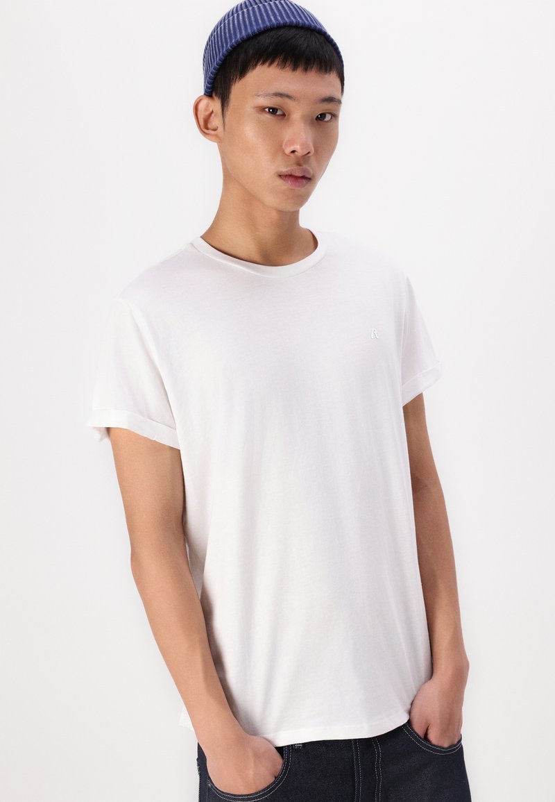 Weißes Baumwoll-T-Shirt mit umgeschlagenen Ärmeln, runder Halslinie und kleinem gesticktem Logo auf der Brust, kombiniert mit dunklen Jeans.