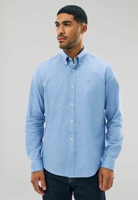 Šviesiai mėlyna marškinėlių palaidinė su apykakle, ilgomis rankovėmis ir pritaikytu siluetu. Yra mažas, siuvinėtas logotipas ant krūtinės. Pagaminta iš medvilnės.