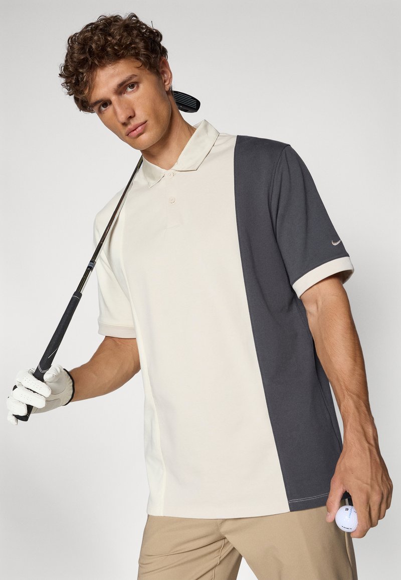 Camisa polo de Nike en color crema y gris oscuro, de manga corta, con diseño bicolor, con cuello, de tela suave, usada con un palo de golf y una pelota.