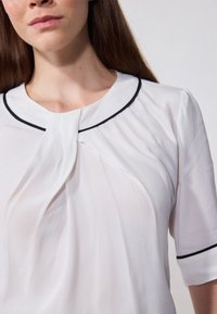 Blouse blanche avec un col structuré et une bordure noire, des manches courtes bouffantes et un tissu doux drapé.