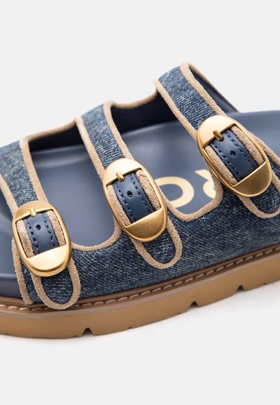 Σανδάλι με τρεις ρυθμιζόμενες χρυσές αγκράφες σε μπλε λουριά από τζιν και ταμπά επένδυση, με χοντρή καφέ σόλα και μπλε εσωτερική σόλα.