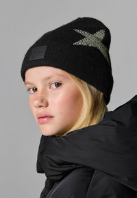 Černá pletená čepice s designem stříbrné hvězdy a koženou nášivkou s logem. Materiál vypadá měkce a má texturu. Model má na sobě černou bundu.