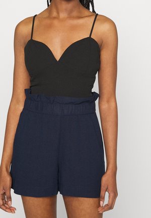 Vrouw met een zwarte, nauwsluitende top met spaghettibandjes, gecombineerd met een high-waisted navyblauwe paperbag-short, op een neutrale achtergrond.