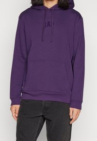 Sudadera morada con bolsillo frontal, capucha ajustable con cordón y logotipo en relieve, hecha de una tela suave con una textura lisa.