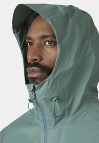 Detailní záběr muže s vousy, který má na sobě světle zelenou pláštěnku s kapucí staženou kolem obličeje.