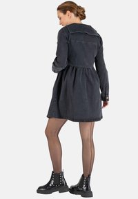Schwarzes Denim-Kleid mit langen Ärmeln, tailliertem Oberteil und auslaufendem Rock. Mit einem knopfbaren Kragen und strukturiertem Stoff ausgestattet. Akzentuiert mit gemusterten Strumpfhosen und glänzenden schwarzen Ankle Boots.