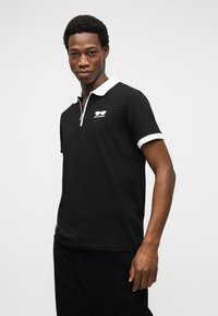 Άνδρας που φοράει μαύρο πόλο με κοντά μανίκια, λευκή γιακά και λεπτομέρεια στα μανίκια, στέκεται μπροστά σε απλό ανοιχτό γκρι φόντο.