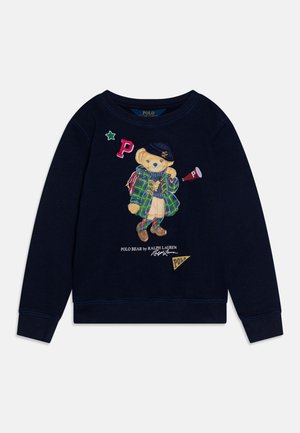 Granatowy sweter z haftowanym niedźwiedziem w kratkowanej kurtce, berecie i mokasynach, otoczony literami i logo na łatkach.