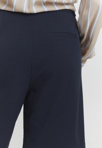 Námořnické modré kalhoty s hladkou texturou, s jemnou zadní kapsou a vysokým pasem, v kombinaci s béžovou pruhovanou blůzou.