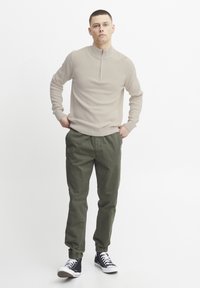 Pull beige texturé avec une fermeture éclair quart, associé à un pantalon cargo vert. Le mannequin porte des baskets montantes noires. L'arrière-plan est blanc uni.