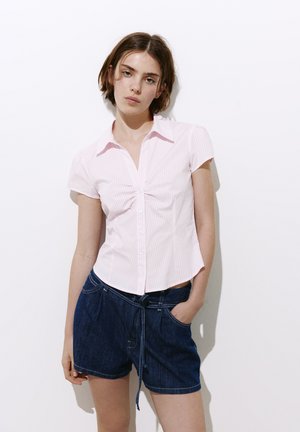 Jeune femme aux cheveux bruns courts, portant un t-shirt rose clair à manches courtes et un short en jean bleu foncé, debout contre un mur blanc.