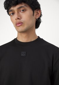 Svart hettegenser med rund hals, laget av et glatt stoff. Har en svart gummilogo-patch på brystet.