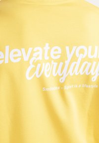 Żółta bawełniana koszulka z dużym białym napisem, który głosi "Podnieś swoją codzienność" oraz "Smilodox - sport to styl życia." Miękka w dotyku.