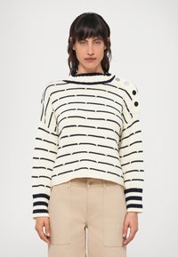 Kreminė megztinė su tamsiai mėlynomis juostelėmis, apvaliu iškirpimu ir mygtukų akcentais ant peties. Megzto audinio struktūra, laisvas kirpimas. Derinama su šviesiai rudos spalvos kelnėmis.