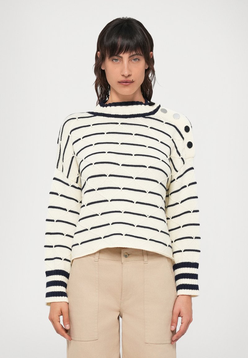 Kreminė megztinė su tamsiai mėlynomis juostelėmis, apvaliu iškirpimu ir mygtukų akcentais ant peties. Megzto audinio struktūra, laisvas kirpimas. Derinama su šviesiai rudos spalvos kelnėmis.