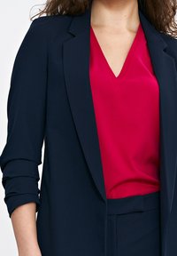 Granatowa marynarka o zwartym kroju, z podwiniętymi rękawami. Noszona na różowej bluzce z dekoltem w serek, podkreślając warstwowy styl.