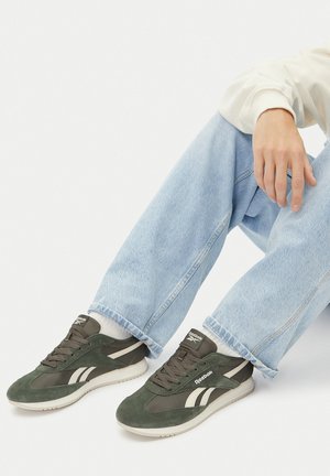 Zielone zamszowe trampki z białymi akcentami, zapinane na sznurowadła, z gumową podeszwą. Noszone z jasnoniebieskimi dżinsowymi spodniami i białymi skarpetkami.